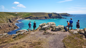 Looking towards Nanjizal This landscape photograph shows several people standing and sitting on rocky terrain overlooking the turquoise sea and dramatic cliffs along the coast of Cornwall, England, in the United Kingdom. The main subject is the view towards Nanjizal, with Nanjizal Beach visible below the cliffs. The image was captured in the early afternoon during the summer season, as indicated by the bright sunlight, clear blue sky, and the people dressed in light clothing suitable for warm weather. The rugged cliffs, green coastal headlands, and deep blue water of the sea characterize the area, highlighting the scenic beauty of Cornwall and the natural appeal of this section of the coastline near Nanjizal Beach.